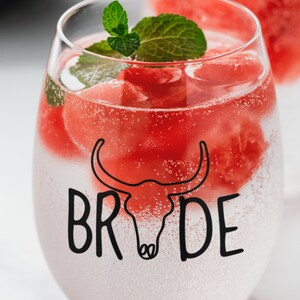May include: A clear glass with a watermelon-themed design. The glass is filled with watermelon cubes, sparkling water, and a sprig of mint. The text "BRIDE" is printed on the glass.