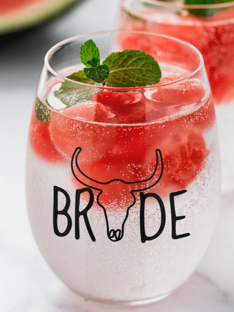 May include: A clear glass with a watermelon-themed design. The glass is filled with watermelon cubes, sparkling water, and a sprig of mint. The text "BRIDE" is printed on the glass.