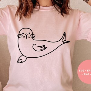 May include: A pink t-shirt with a black line drawing of a seal on the front. The seal is facing left and has a simple, cartoon-like design.