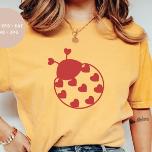 Könnte beinhalten: Ein gelbes T-Shirt mit einem roten Marienkäfer-Design mit einem Herzmuster. Der Marienkäfer hat einen roten Körper und schwarze Punkte. Das Design ist mittig auf dem Shirt platziert.
