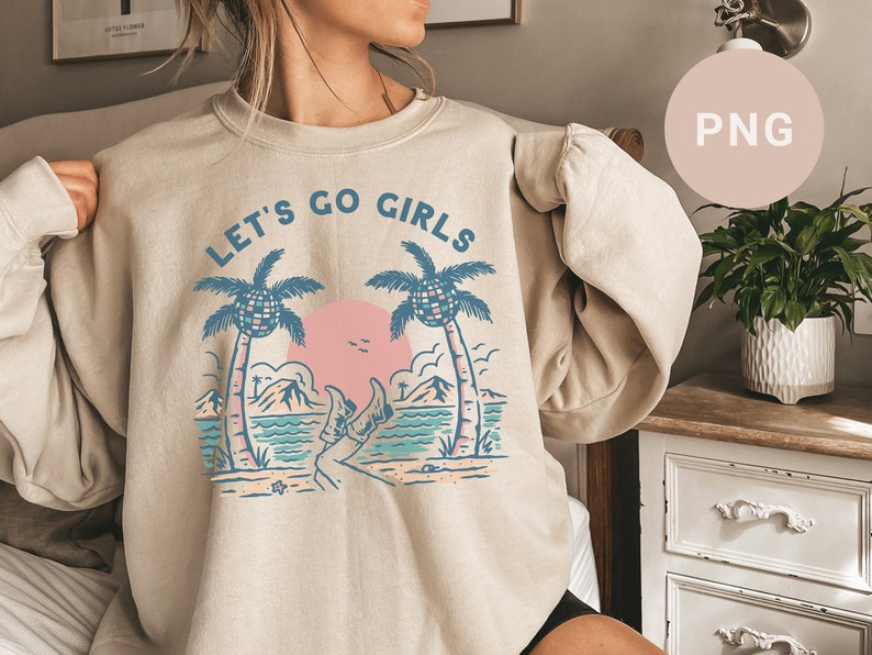 K&ouml;nnte beinhalten: Beiges Sweatshirt mit tropischem Aufdruck. Das Design zeigt zwei Palmen, eine rosa Sonne und den Text "LET'S GO GIRLS" in Blau. Die Grafik enth&auml;lt auch eine Strandszene mit Bergen und einer Person mit Stiefeln.