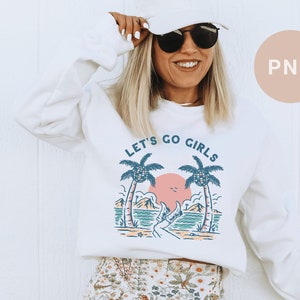 K&ouml;nnte beinhalten: Wei&szlig;es Sweatshirt mit dem Text "LET'S GO GIRLS" &uuml;ber einer Strandszene mit Palmen, einer rosa Sonne und t&uuml;rkisfarbenem Wasser. Das Sweatshirt hat einen Rundhalsausschnitt und lange &Auml;rmel.