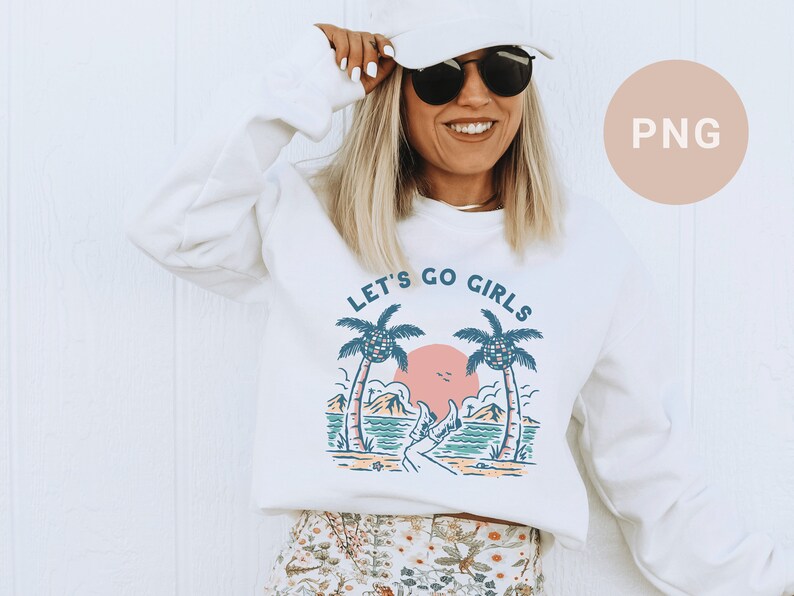 K&ouml;nnte beinhalten: Wei&szlig;es Sweatshirt mit dem Text "LET'S GO GIRLS" &uuml;ber einer Strandszene mit Palmen, einer rosa Sonne und t&uuml;rkisfarbenem Wasser. Das Sweatshirt hat einen Rundhalsausschnitt und lange &Auml;rmel.