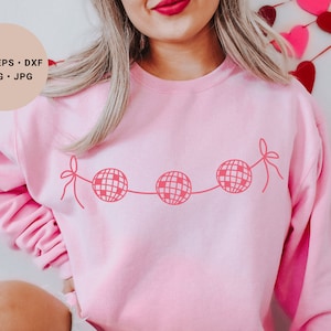 May include: Pink sweatshirt with a graphic of a string of disco balls with red bows.