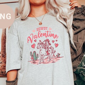 May include: A grey t-shirt with a pink and white graphic design featuring a cowgirl riding a horse with the text "Howdy Valentine".