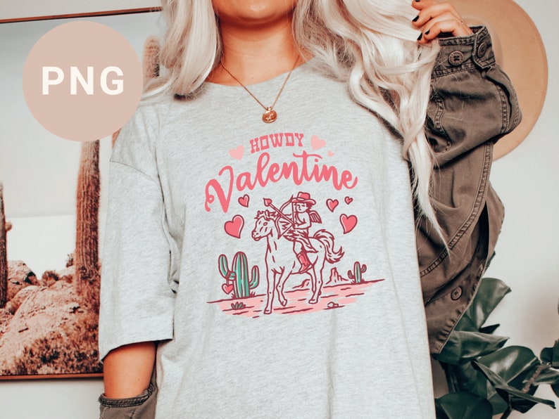 May include: A grey t-shirt with a pink and white graphic design featuring a cowgirl riding a horse with the text "Howdy Valentine".