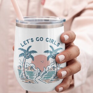 K&ouml;nnte beinhalten: Wei&szlig;er isolierter Weinglasbecher mit Deckel und rosa Strohhalm. Der Becher zeigt eine Strandszene mit Palmen, einem Sonnenuntergang und dem Text "LET'S GO GIRLS" in Blau. Eine Hand h&auml;lt den Becher.