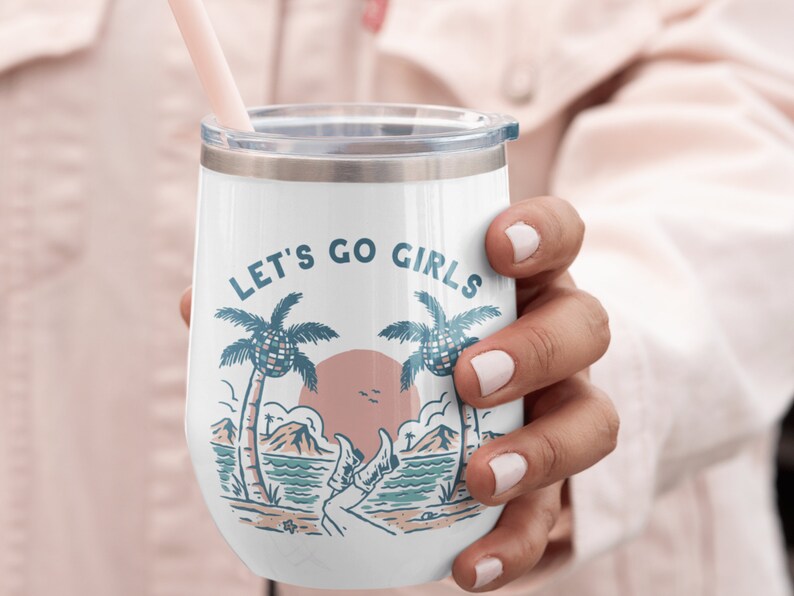 K&ouml;nnte beinhalten: Wei&szlig;er isolierter Weinglasbecher mit Deckel und rosa Strohhalm. Der Becher zeigt eine Strandszene mit Palmen, einem Sonnenuntergang und dem Text "LET'S GO GIRLS" in Blau. Eine Hand h&auml;lt den Becher.