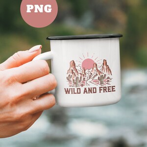 Puede incluir: Taza de esmalte blanco con borde negro. La taza presenta un gr&aacute;fico de un paisaje des&eacute;rtico con un sol rojo, monta&ntilde;as y cactus. El texto "WILD AND FREE" est&aacute; impreso en marr&oacute;n debajo del gr&aacute;fico.