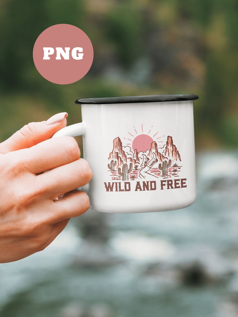 Puede incluir: Taza de esmalte blanco con borde negro. La taza presenta un gr&aacute;fico de un paisaje des&eacute;rtico con un sol rojo, monta&ntilde;as y cactus. El texto "WILD AND FREE" est&aacute; impreso en marr&oacute;n debajo del gr&aacute;fico.