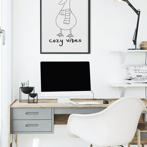 K&ouml;nnte beinhalten: Schwarz-wei&szlig;-Linienkunst-Druck eines Entchens mit Hut und Schal mit dem Text "cozy vibes" darunter. Der Druck ist gerahmt und h&auml;ngt an einer wei&szlig;en Wand &uuml;ber einem Schreibtisch.