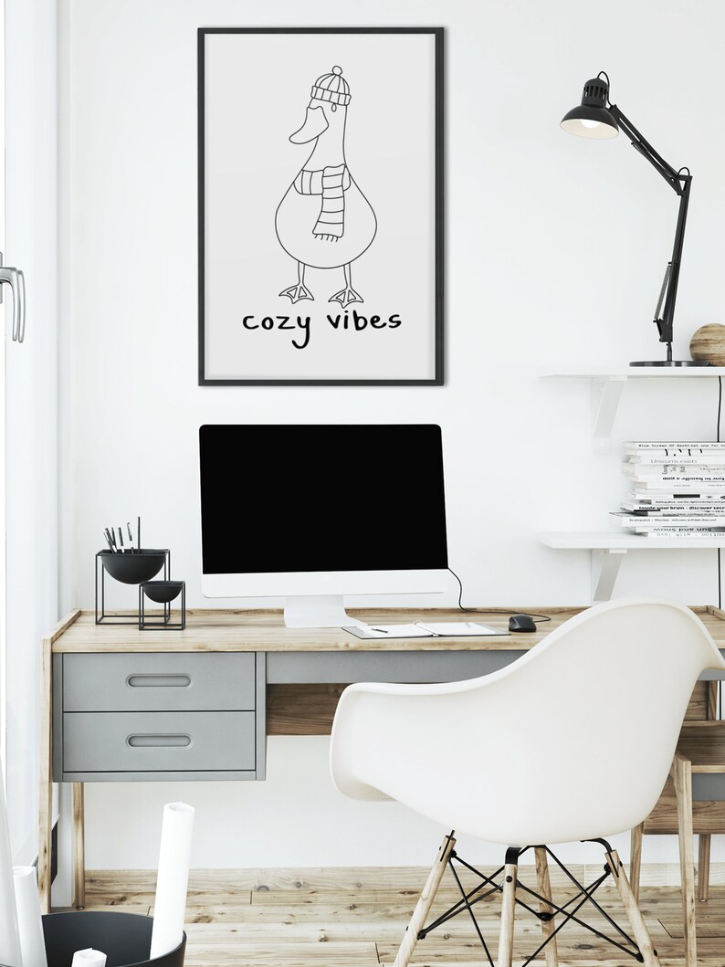 K&ouml;nnte beinhalten: Schwarz-wei&szlig;-Linienkunst-Druck eines Entchens mit Hut und Schal mit dem Text "cozy vibes" darunter. Der Druck ist gerahmt und h&auml;ngt an einer wei&szlig;en Wand &uuml;ber einem Schreibtisch.
