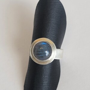 May include: A silver ring featuring a large, round, blue-grey gemstone. The gemstone is set in a silver bezel on a simple silver band. The ring is displayed on a black, curved stand against a white backdrop.