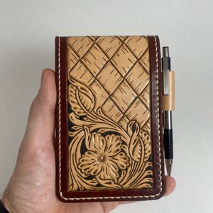 May include: A brown leather notebook cover with a tooled design of a flower and leaves. The cover has a white stitching around the edges and a pen is tucked into the cover.