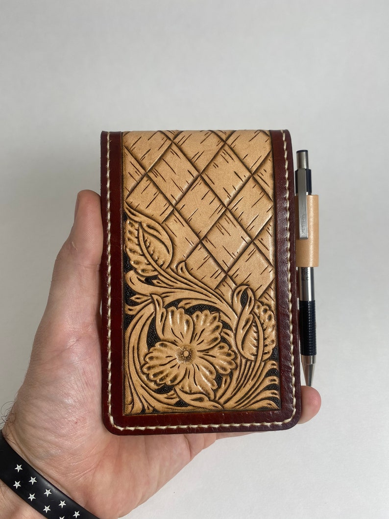 May include: A brown leather notebook cover with a tooled design of a flower and leaves. The cover has a white stitching around the edges and a pen is tucked into the cover.