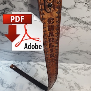 Può includere: Un cinturino in pelle marrone con il nome "CHARLIE" inciso su di esso. Il cinturino ha un motivo decorativo ed è progettato per una chitarra.