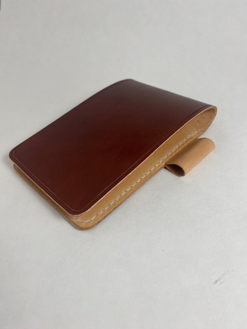May include: A brown leather wallet with a tan leather flap and stitching. The wallet is closed and lying on a white surface.