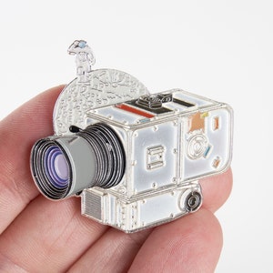 Puede incluir: Un pin de esmalte plateado que representa una cámara de estilo vintage con una lente, una superficie lunar y una pequeña figura de astronauta. La cámara tiene un cuerpo blanco con detalles negros y una lente plateada.