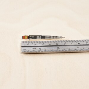K&ouml;nnte beinhalten: Eine silberne Emaille-Anstecknadel mit einer Rakete mit orangefarbenen Flammen. Die Nadel ist etwa 5 cm lang und wird mit einem Lineal gemessen.