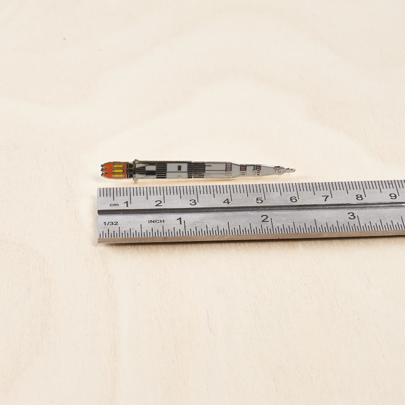 K&ouml;nnte beinhalten: Eine silberne Emaille-Anstecknadel mit einer Rakete mit orangefarbenen Flammen. Die Nadel ist etwa 5 cm lang und wird mit einem Lineal gemessen.
