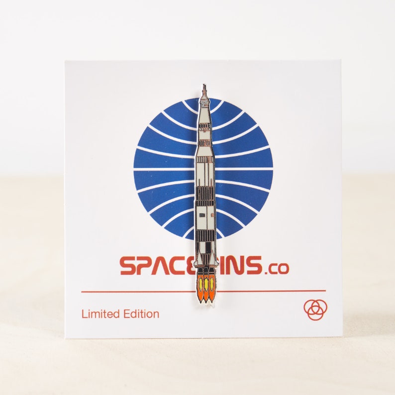 K&ouml;nnte beinhalten: Eine wei&szlig;e und orangefarbene Emaille-Anstecknadel mit einer startenden Rakete vor einem blauen und wei&szlig;en Kreis als Hintergrund. Die Anstecknadel tr&auml;gt die Aufschrift "Space Ins.co" und "Limited Edition".