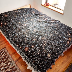 Op de afbeelding: Een zwarte deken met een galaxy print met een wit, zilver en gouden sterrenpatroon. De deken is over een bed gelegd met een wit laken en een houten hoofdeinde.