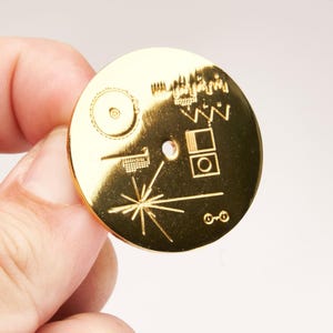 May include: A gold-colored, circular pendant featuring an engraved design. The design includes geometric shapes, lines, and a starburst pattern. The pendant is held in a person's hand.