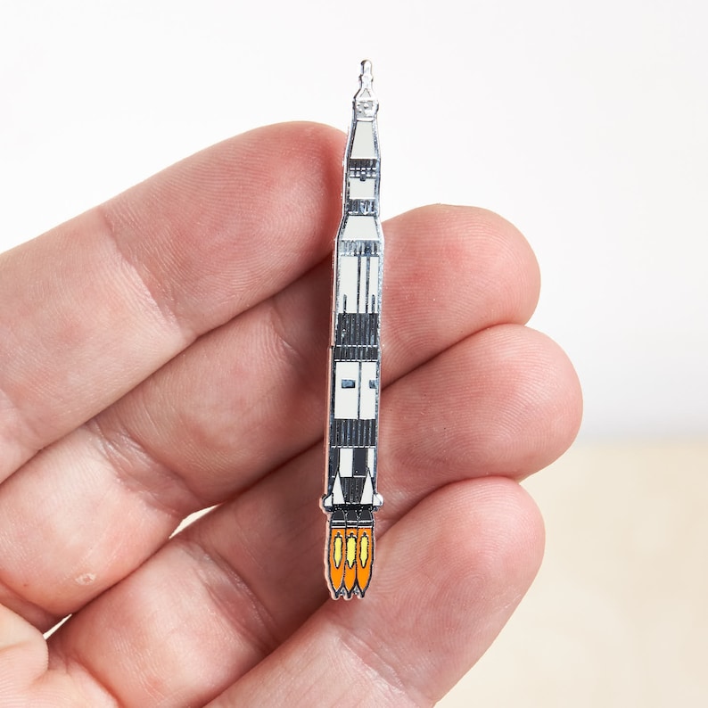 K&ouml;nnte beinhalten: Eine silberne Emaille-Anstecknadel mit einer Rakete mit orangefarbenen Flammen. Die Rakete ist schwarz-wei&szlig; mit silbernem Umriss.
