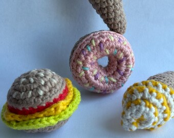 May include: A collection of handmade crochet food items, including a burger, a pink donut with sprinkles, and two ice cream cones. The ice cream cones feature brown and white, and yellow and white colors.