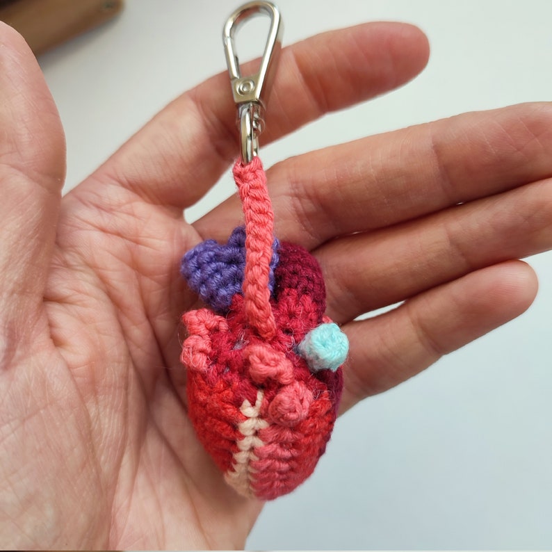 May include: A crocheted heart keyring with a silver key ring. The heart is made of red, pink and purple yarn and has a small blue detail.