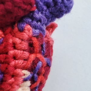May include: Close-up of a crocheted heart-shaped object made with red, pink, and purple yarn. The yarn is woven together in a textured pattern.