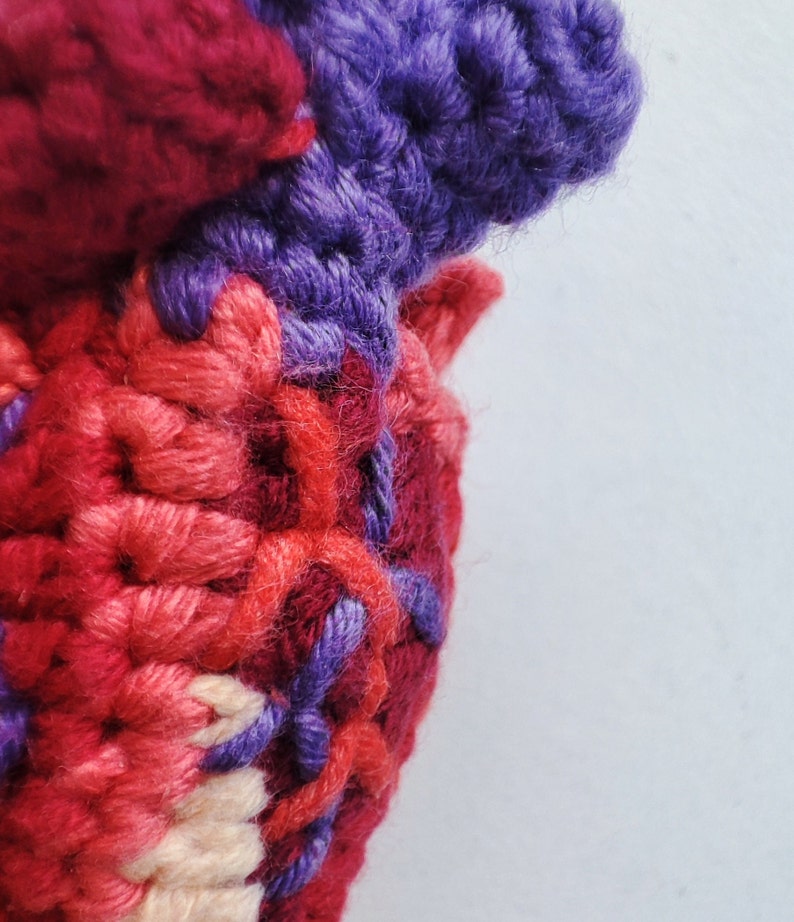 May include: Close-up of a crocheted heart-shaped object made with red, pink, and purple yarn. The yarn is woven together in a textured pattern.