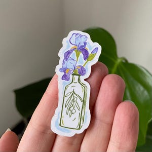Könnte beinhalten: Ein Aufkleber mit einer handgezeichneten Illustration einer durchsichtigen Glasflasche mit einem violetten Irisblumenstrauß darin. Die Flasche enthält einen grünen Olivenzweig. Der Aufkleber hat einen weißen Hintergrund.
