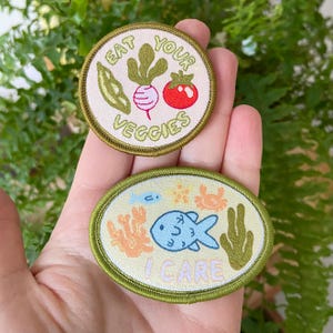 Puede incluir: Dos parches bordados para planchar. Un parche es verde y blanco con el texto "Eat Your Veggies" y una imagen de dibujos animados de un tomate, un rábano y una vaina de guisante. El otro parche es verde y amarillo con el texto "I Care" y una imagen de dibujos animados de un pez, coral y un cangrejo.