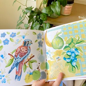 K&ouml;nnte beinhalten: Offenes Buch mit Aquarellillustrationen. Eine Seite zeigt einen Vogel auf einem Ast mit blauen Blumen und Beeren. Die andere Seite hat eine Banane, eine Birne, Blaubeeren und den Text "WHAT DID YOU DO TODAY?"