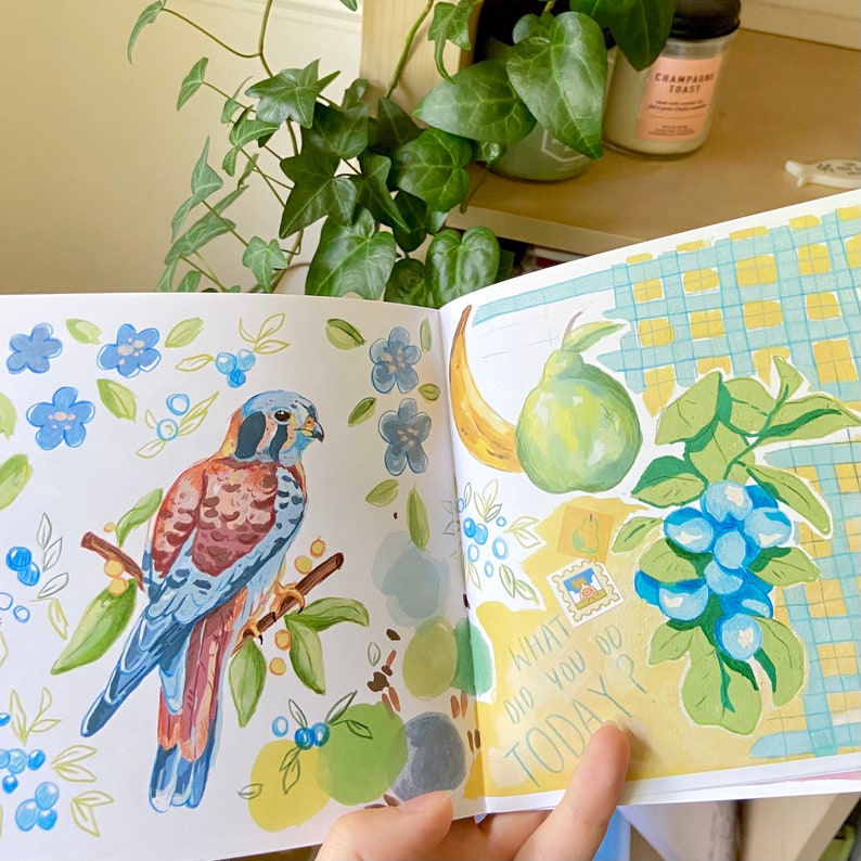 K&ouml;nnte beinhalten: Offenes Buch mit Aquarellillustrationen. Eine Seite zeigt einen Vogel auf einem Ast mit blauen Blumen und Beeren. Die andere Seite hat eine Banane, eine Birne, Blaubeeren und den Text "WHAT DID YOU DO TODAY?"