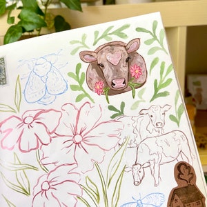 K&ouml;nnte beinhalten: Aquarellillustration mit einer braunen Kuh mit herzf&ouml;rmiger Markierung, umgeben von rosa Blumen und gr&uuml;nen Bl&auml;ttern. Skizzen von Blumen, K&uuml;hen und einer Motte sind ebenfalls vorhanden, was ein skurriles, pastorales Thema ergibt.