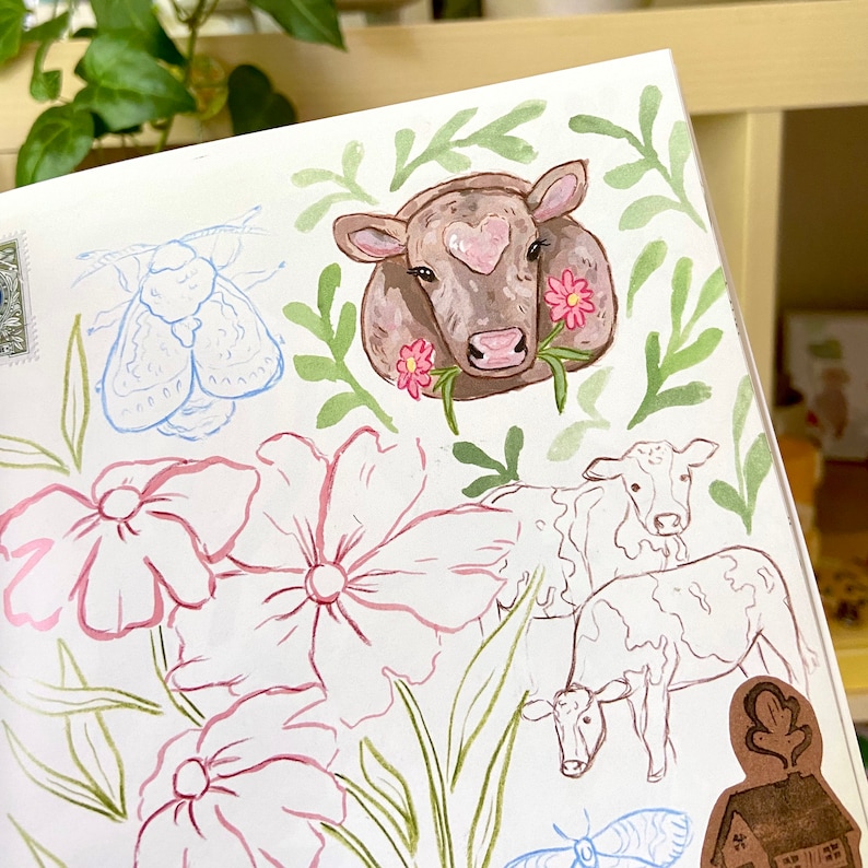 K&ouml;nnte beinhalten: Aquarellillustration mit einer braunen Kuh mit herzf&ouml;rmiger Markierung, umgeben von rosa Blumen und gr&uuml;nen Bl&auml;ttern. Skizzen von Blumen, K&uuml;hen und einer Motte sind ebenfalls vorhanden, was ein skurriles, pastorales Thema ergibt.