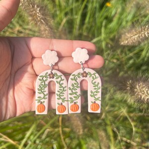 May include: A pair of cream-colored, arch-shaped earrings with a fall theme. Each earring features a pumpkin, green vines, and small brown flowers. The earrings are held up by a light pink cloud-shaped stud.