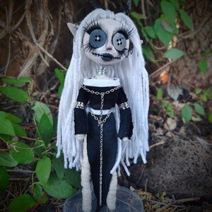 May include: A doll with long white yarn hair, black horns, and large button eyes. It features long eyelashes, a stitched mouth, and a black dress with a white collar. The doll is adorned with silver chain accents and stands on a clear circular base.