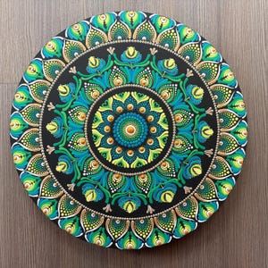 May include: A round, black mandala art piece with a detailed, symmetrical design. The artwork features concentric circles with teal, green, and gold accents. The mandala is approximately 12 inches in diameter.