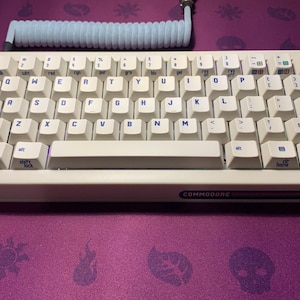 May include: A white Commodore 64 keyboard with blue and purple lettering. The keyboard has a white space bar and a black space bar with the word "COMMODORE" printed on it. The keyboard is connected to a blue coiled cable.