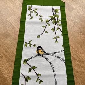 Könnte beinhalten: Ein weißer Tischläufer mit grünem Rand zeigt ein zartes Design aus Birkenzweigen und Blättern mit einem kleinen gelben Vogel, der auf einem Zweig sitzt.
