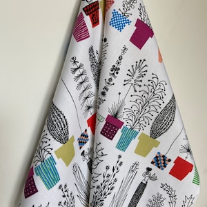 Puede incluir: Paño de cocina blanco con un diseño botánico colorido. El paño presenta dibujos lineales negros de plantas y flores en macetas en varios colores, incluyendo rojo, morado, azul y amarillo. El paño está colgado contra una pared blanca.