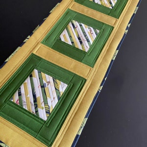 Può includere: Un runner da tavolo rettangolare con un design trapuntato. Il runner presenta tre quadrati verdi con centri in tessuto fantasia, incorniciati da bordi dorati e verdi. I bordi sono rifiniti con un tessuto blu scuro con un motivo verde chiaro.