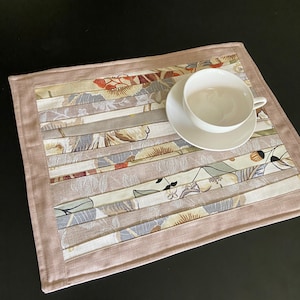 May include: A rectangular placemat with a floral pattern in shades of beige, gray, and red, with a light brown border. A white teacup and saucer sit on the placemat, creating a coordinated table setting.