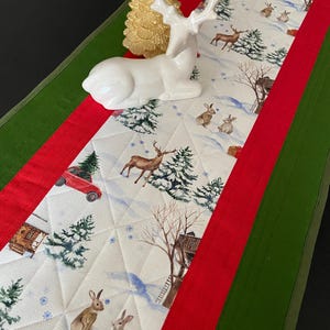 Peut inclure: Un chemin de table matelassé avec un imprimé de forêt hivernale représentant des cerfs, des lapins, des sapins et des voitures rouges sur fond crème, bordé de bandes de tissu rouge et vert. Une figurine de cerf en céramique blanche est posée.