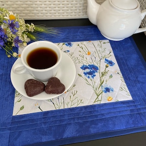 May include: A white teacup and saucer with dark tea and two heart-shaped chocolates sit on a floral placemat. A white teapot is in the background. The placemat has a blue border and a floral pattern with blue flowers.