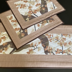 May include: Two rectangular quilted placemats and a matching table runner featuring a floral pattern with white, tan, and dark brown blossoms on a light background. The items have solid tan borders with stitched lines and dark brown edges.