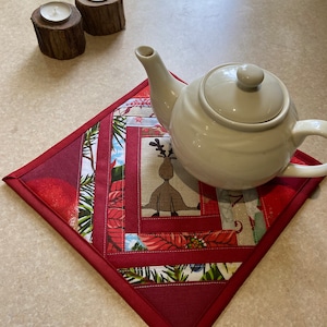Op de afbeelding: Een witte keramische theepot staat op een vierkante gewatteerde stoffen mat met een patchworkontwerp van rode, groene en beige patronen, inclusief een centrale rendierillustratie, vergezeld door twee kleine houten theelichthouders.